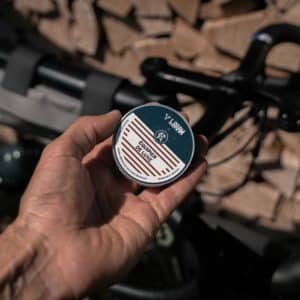 Close-up of a hand holding a tin labeled 'Baume Deluxe' in front of a road bike handlebar and a stacked woodpile.