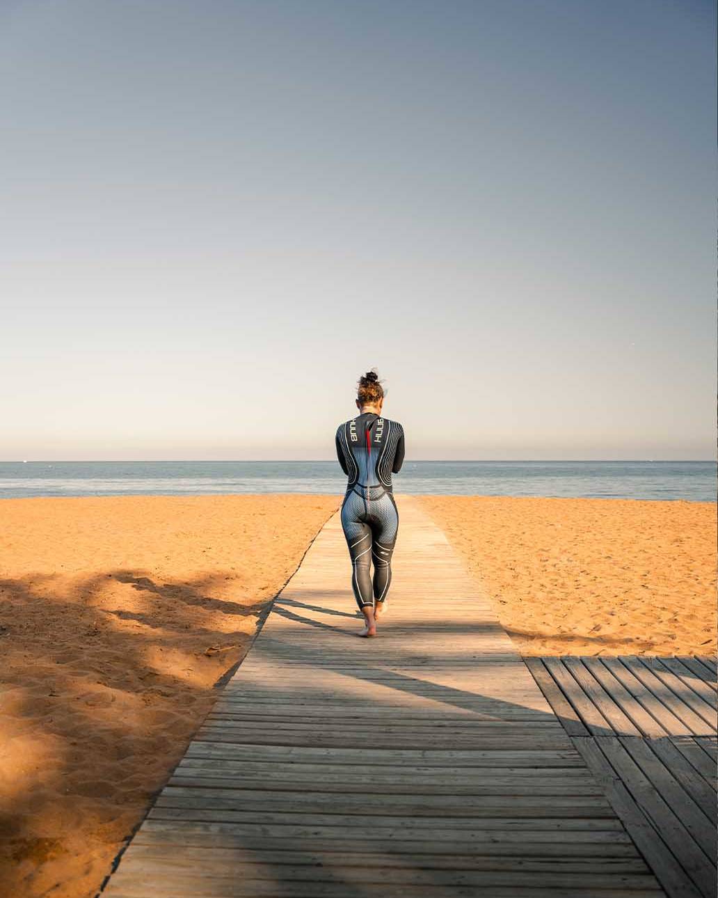 A triathlete in a wetsuit walking along a wooden boardwalk towards a serene sandy beach and calm sea, ideal for triathlon training.