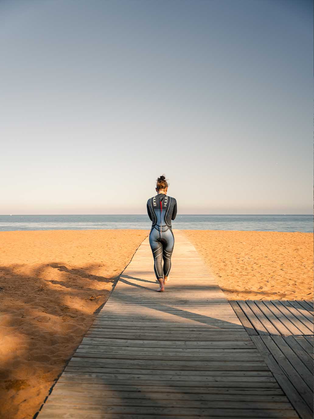 A triathlete in a wetsuit walking along a wooden boardwalk towards a serene sandy beach and calm sea, ideal for triathlon training.