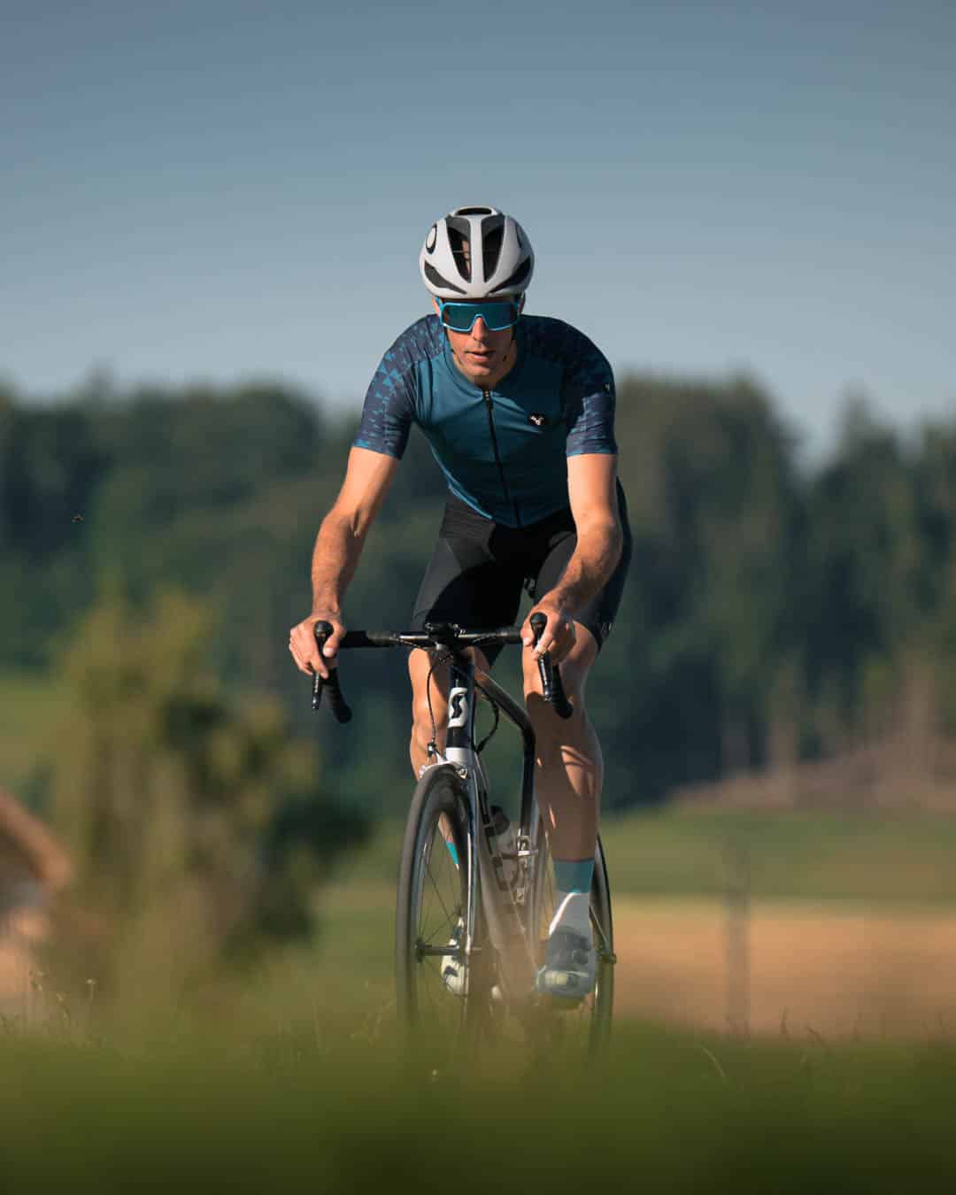 Cyclist riding on a road bike wearing a helmet and sunglasses, set against a blurred green countryside background.