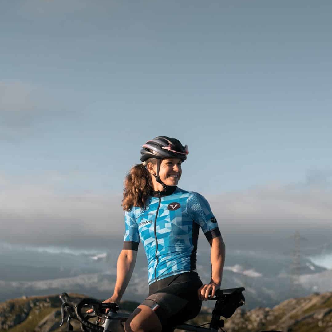 A smiling cyclist in a blue jersey rests on her bike with stunning alpine scenery in the background, exuding joy and determination.