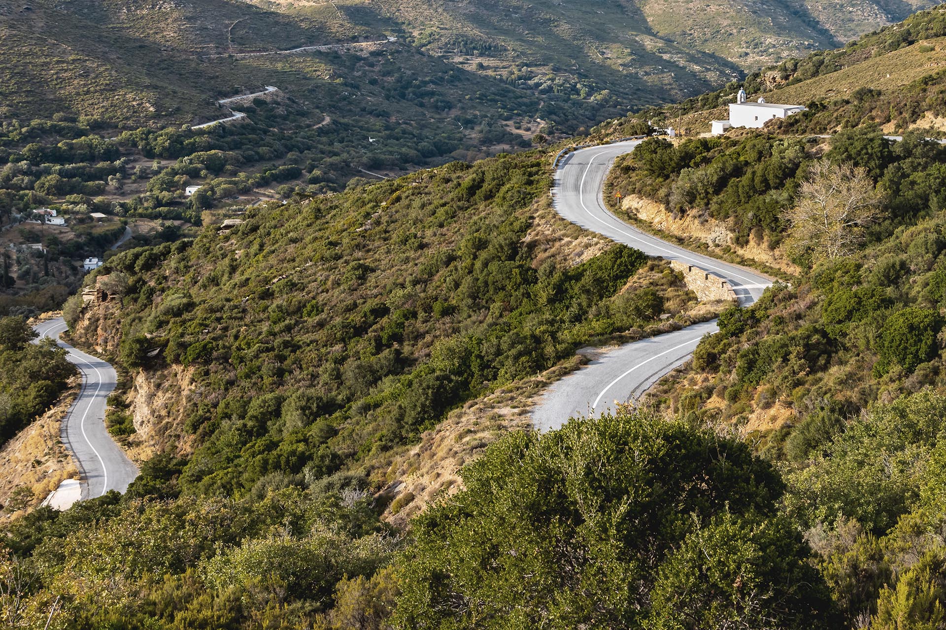 Serpentines of a mountain road with a small church in the highlands of the Greek Cyclades island Andros in the evening light