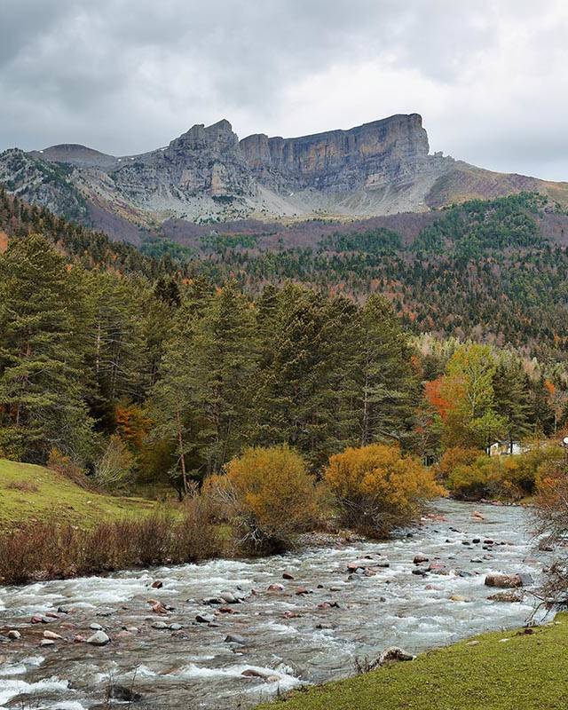 A scenic autumn view of a flowing river, lush forests, and towering cliffs in the Pyrenees, highlighting the stunning routes available for Pyrenees cycling enthusiasts.