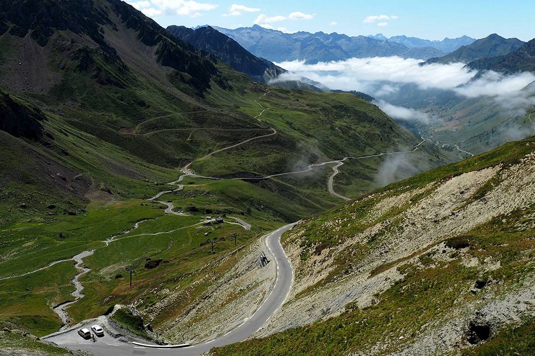 Scenic winding mountain road in the Pyrenees, surrounded by lush green valleys and towering peaks under a clear blue sky.