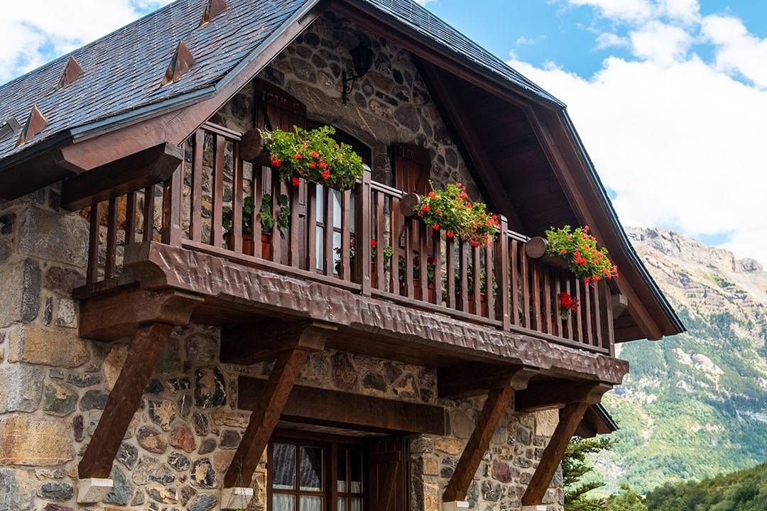 Charming stone chalet with wooden balcony adorned with vibrant flower pots, set against a backdrop of lush green mountains and blue sky.