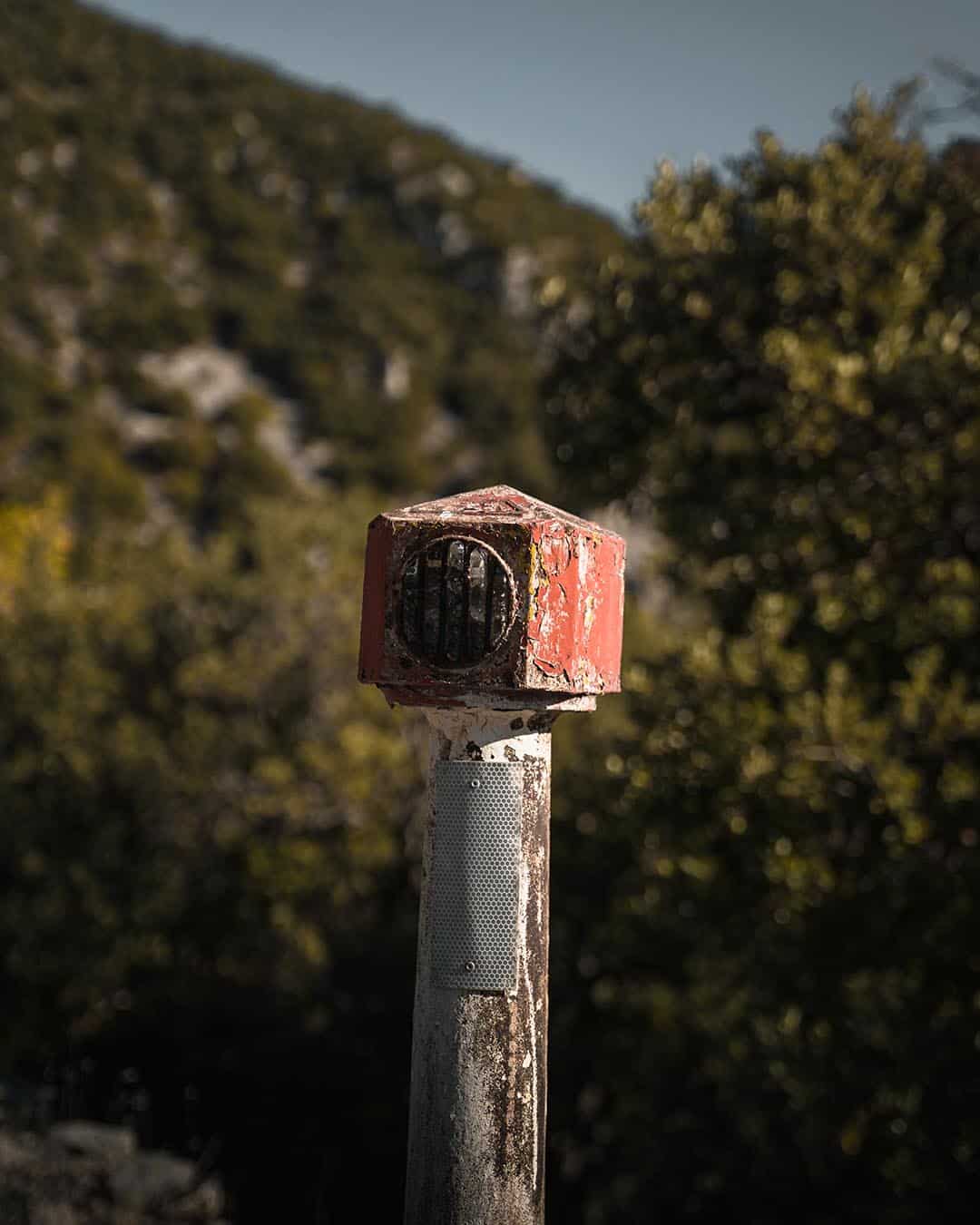 Rustic red metal road marker in a scenic wooded landscape on a sunny day.