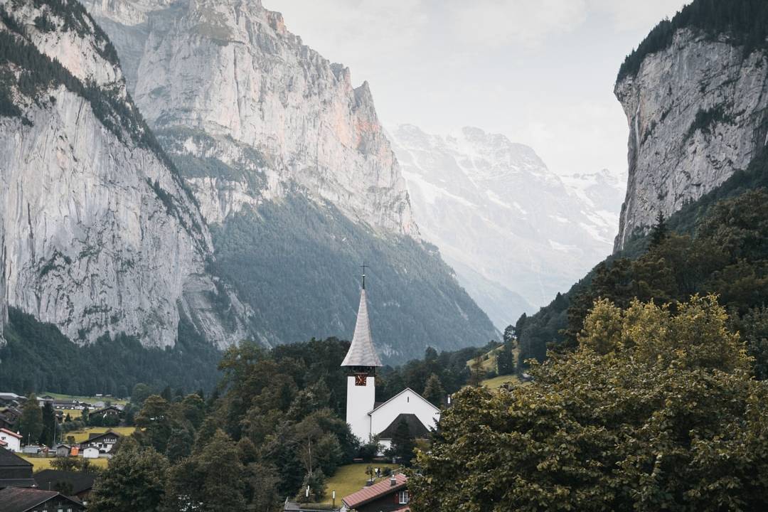 A picturesque Swiss village nestled in a lush green valley, surrounded by towering cliffs and snow-capped mountains, with a charming white church at the center.