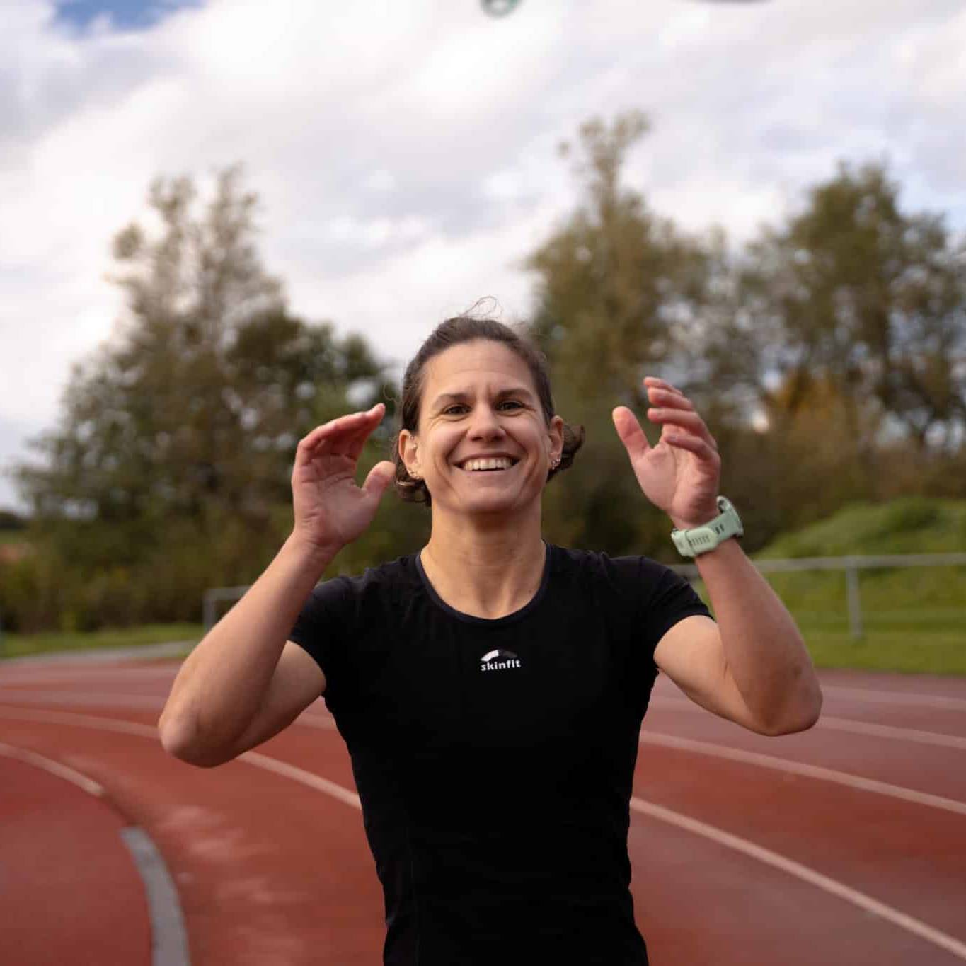 An athlete in a black sports outfit smiles vibrantly on a track, with her hands raised mid-action against a background of trees and a cloudy sky.