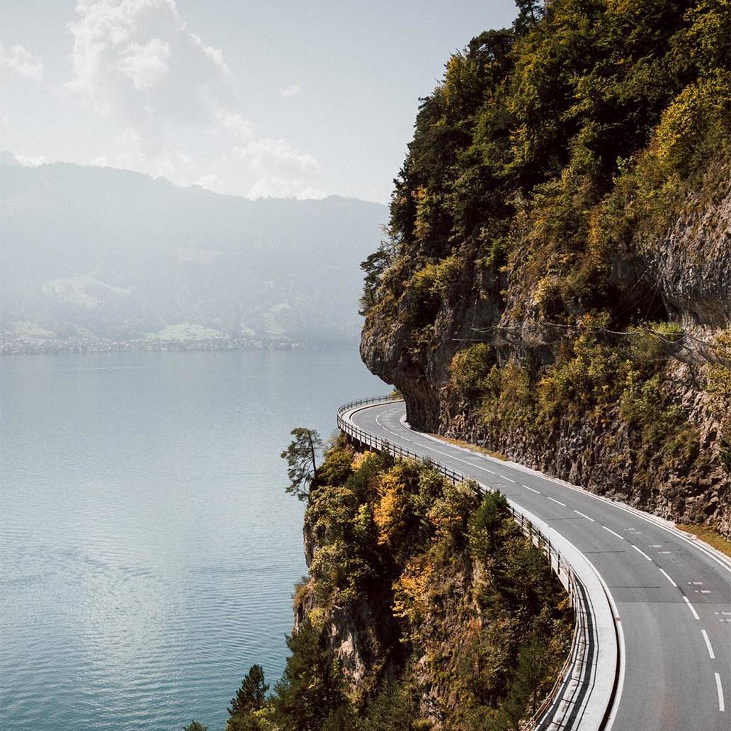 Scenic road curving along a cliffside above a serene lake, surrounded by lush greenery and distant mountains under a clear sky in Switzerland.