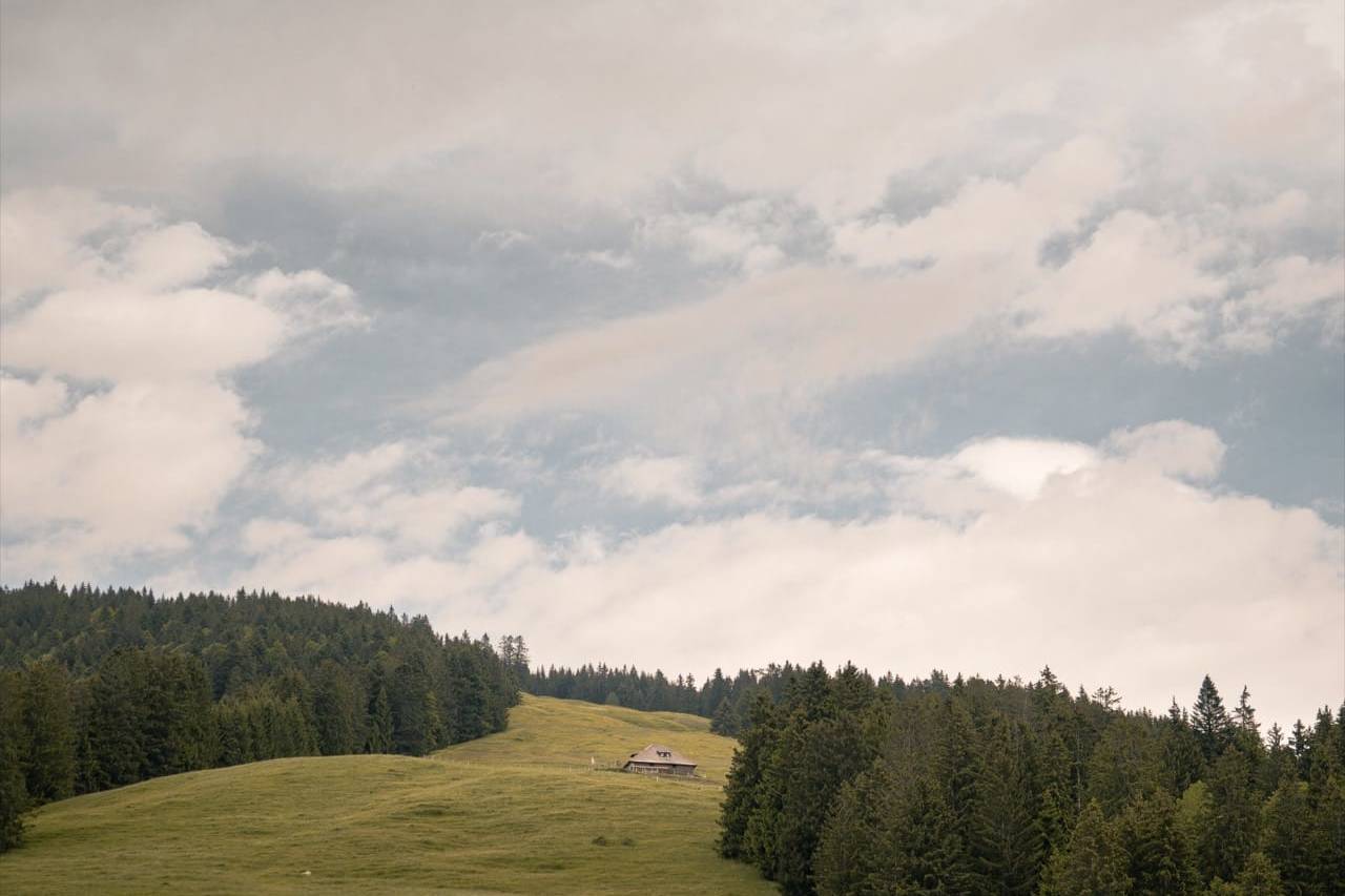 Peaceful Swiss countryside with a solitary cabin on a grassy hill, surrounded by lush green forest under a vast, cloud-filled sky.