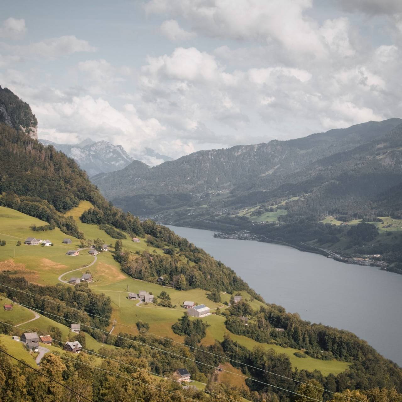 Scenic view of Swiss countryside with small houses scattered across green hills, overlooking a tranquil lake and surrounded by mountainous terrain under a partly cloudy sky.