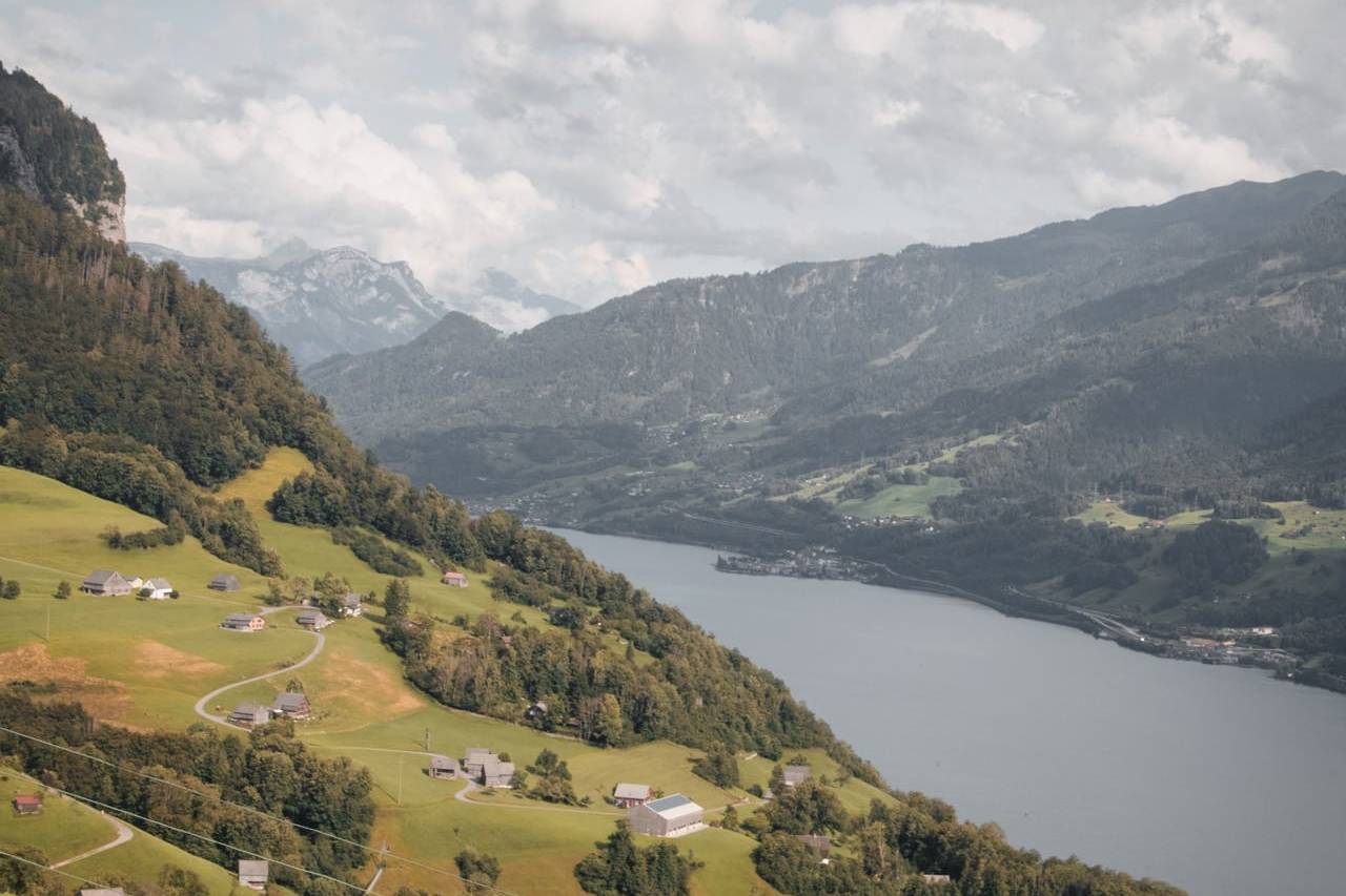 Scenic view of Swiss countryside with small houses scattered across green hills, overlooking a tranquil lake and surrounded by mountainous terrain under a partly cloudy sky.