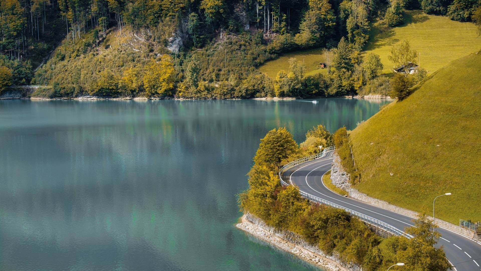 Curving road by a serene turquoise lake in Switzerland, surrounded by lush green hills and dense forested areas with autumn foliage.