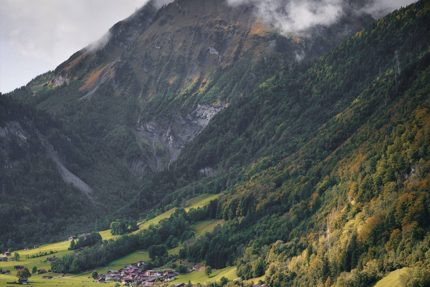 Scenic mountain village in Switzerland with quaint houses nestled between lush green slopes and a calm lake, with misty clouds surrounding the towering mountains above.