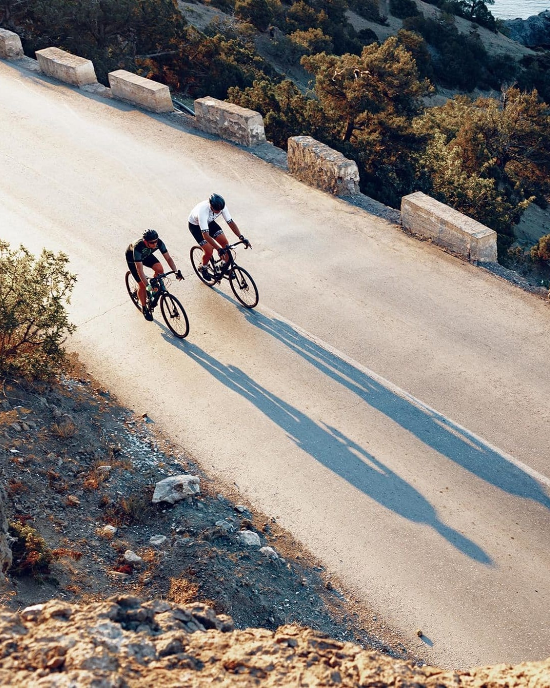 Two cyclists riding side by side on a scenic coastal road, casting long shadows during the golden hour, epitomizing the blend of camaraderie and breathtaking landscapes.