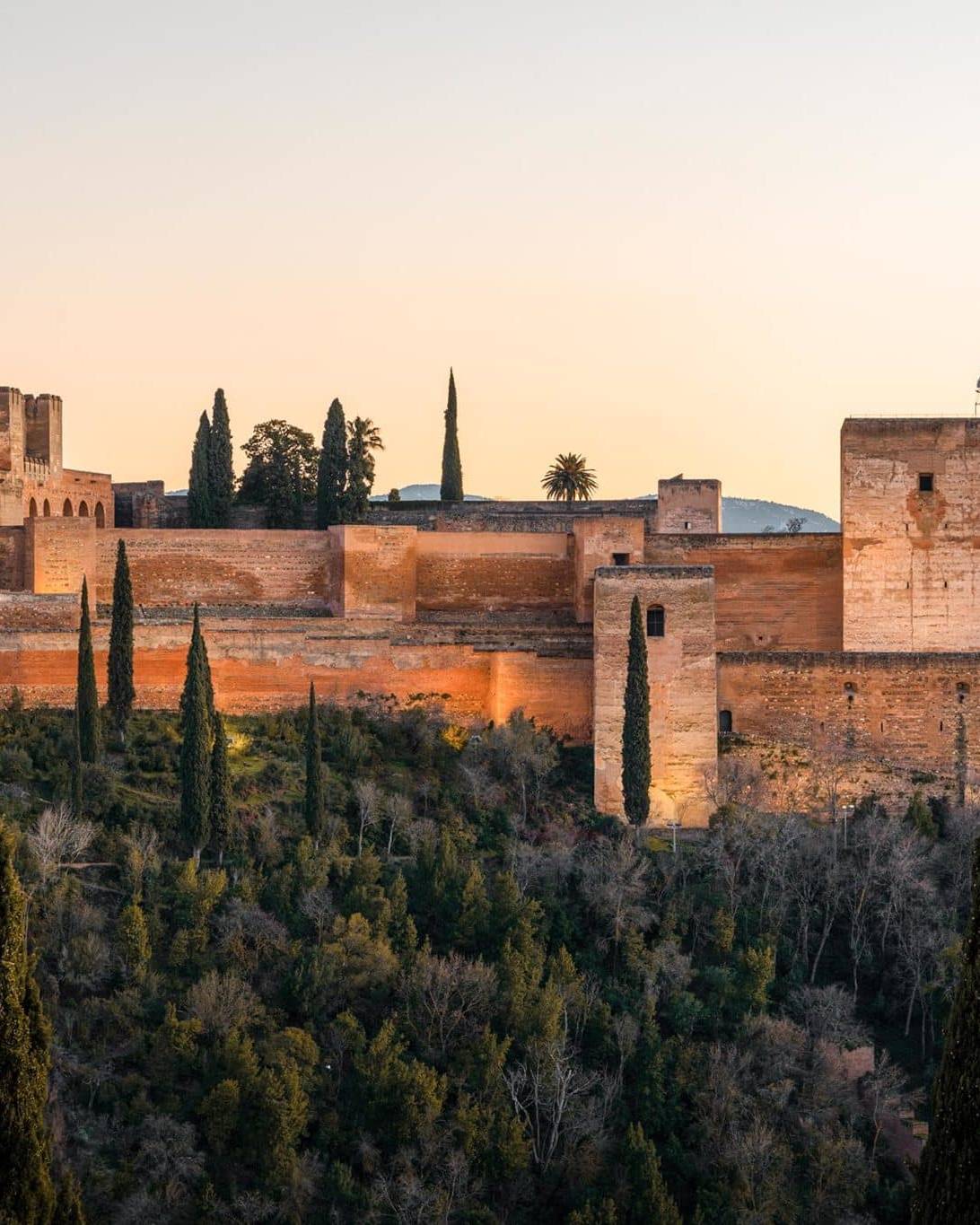 The Alhambra illuminated at sunset with rolling hills and snowy mountains in the background, a must-see landmark for those cycling Andalusia and exploring its rich cultural heritage.