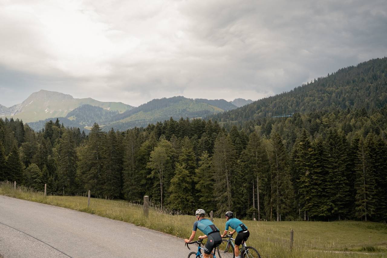 Two cyclists riding up a scenic mountain road, surrounded by lush green forests and distant hills under a cloudy sky, showcasing the tranquility of the Swiss countryside.