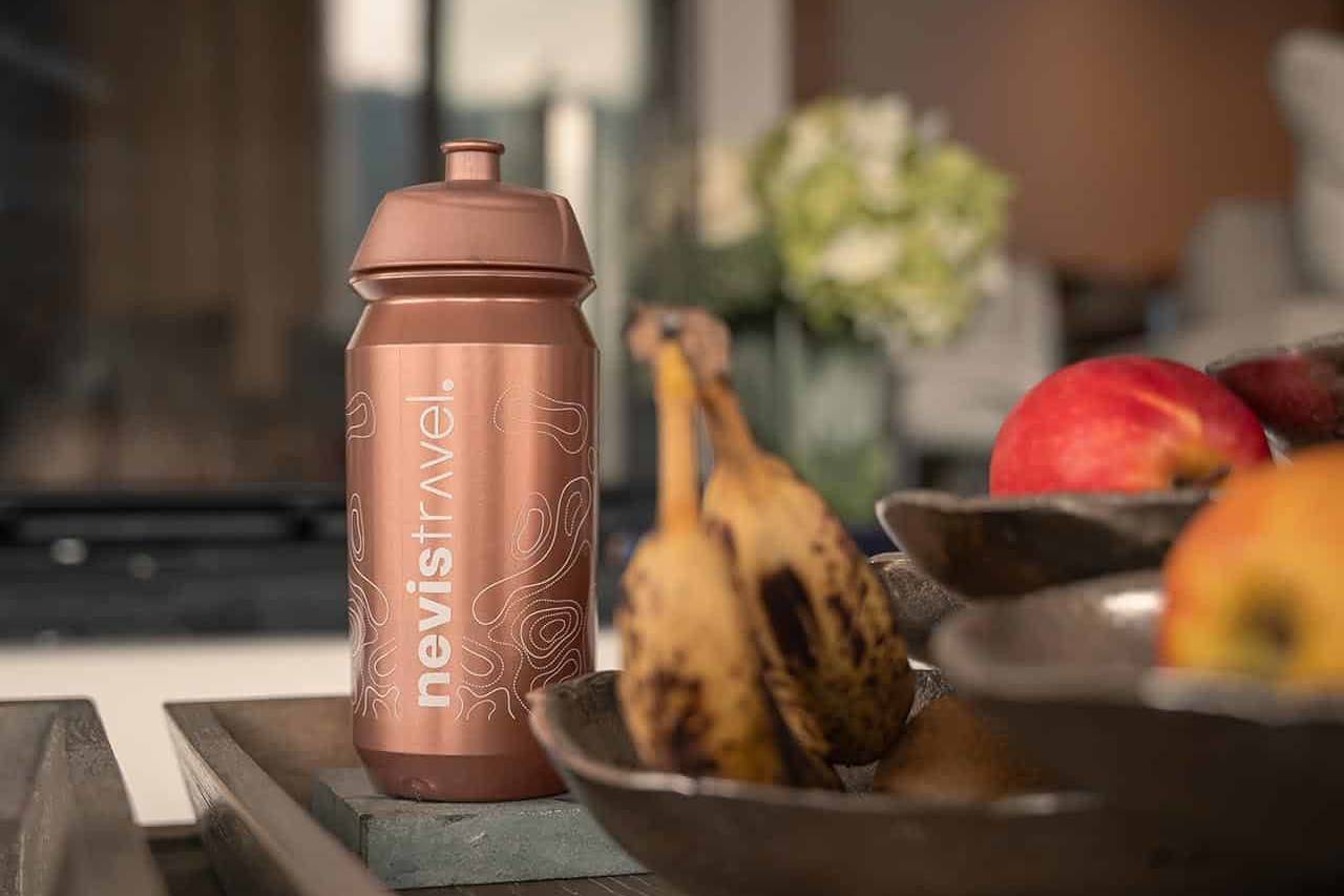 nevistravel-branded water bottle in copper color placed on a wooden tray alongside fresh bananas and apples, creating a warm and inviting atmosphere with a blurred background of indoor decor.