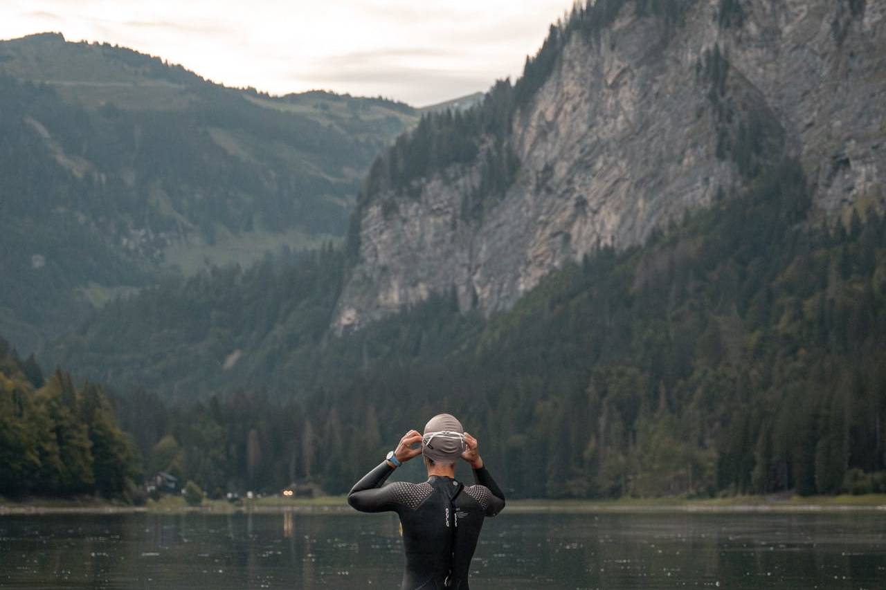 Swimmer in a wetsuit standing waist-deep in a calm lake, adjusting their swim cap, with a mountainous, forested backdrop under a cloudy sky.