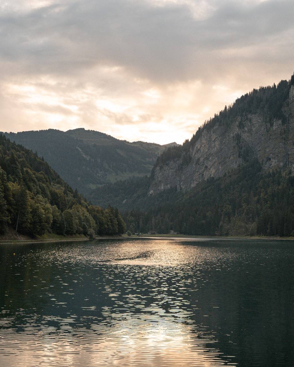 Tranquil lake in Morzine surrounded by forested mountains at sunset, with the sun’s reflection glistening on the water’s surface.