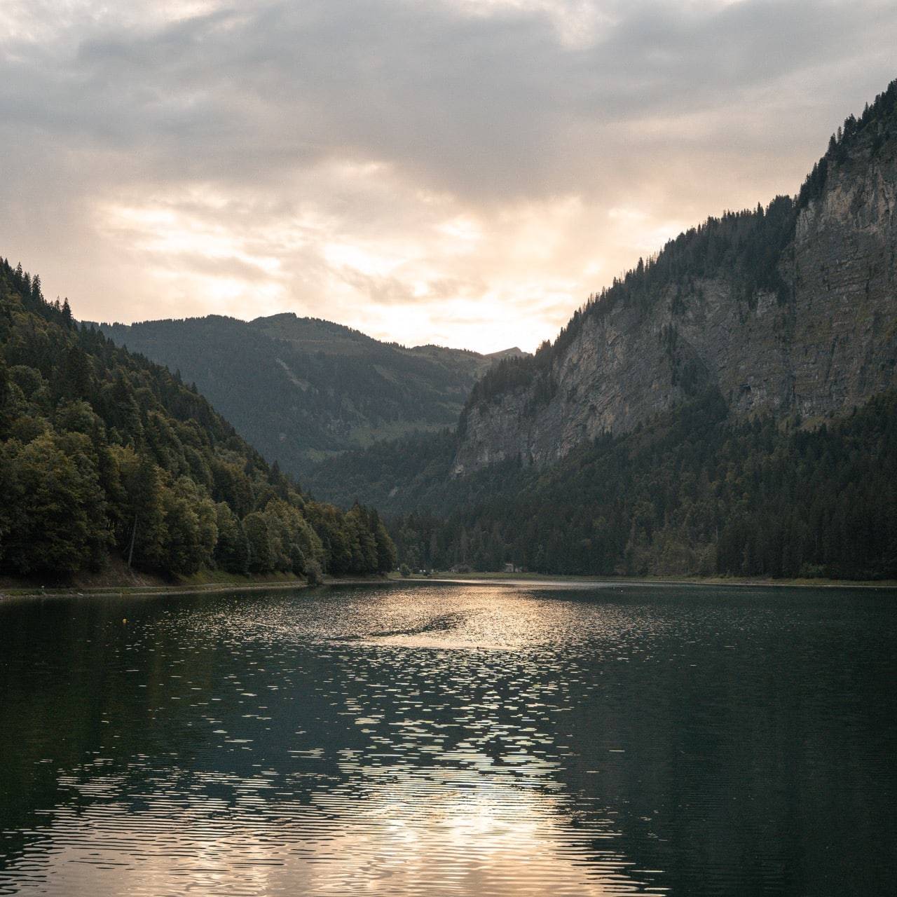 Tranquil lake in Morzine surrounded by forested mountains at sunset, with the sun’s reflection glistening on the water’s surface.