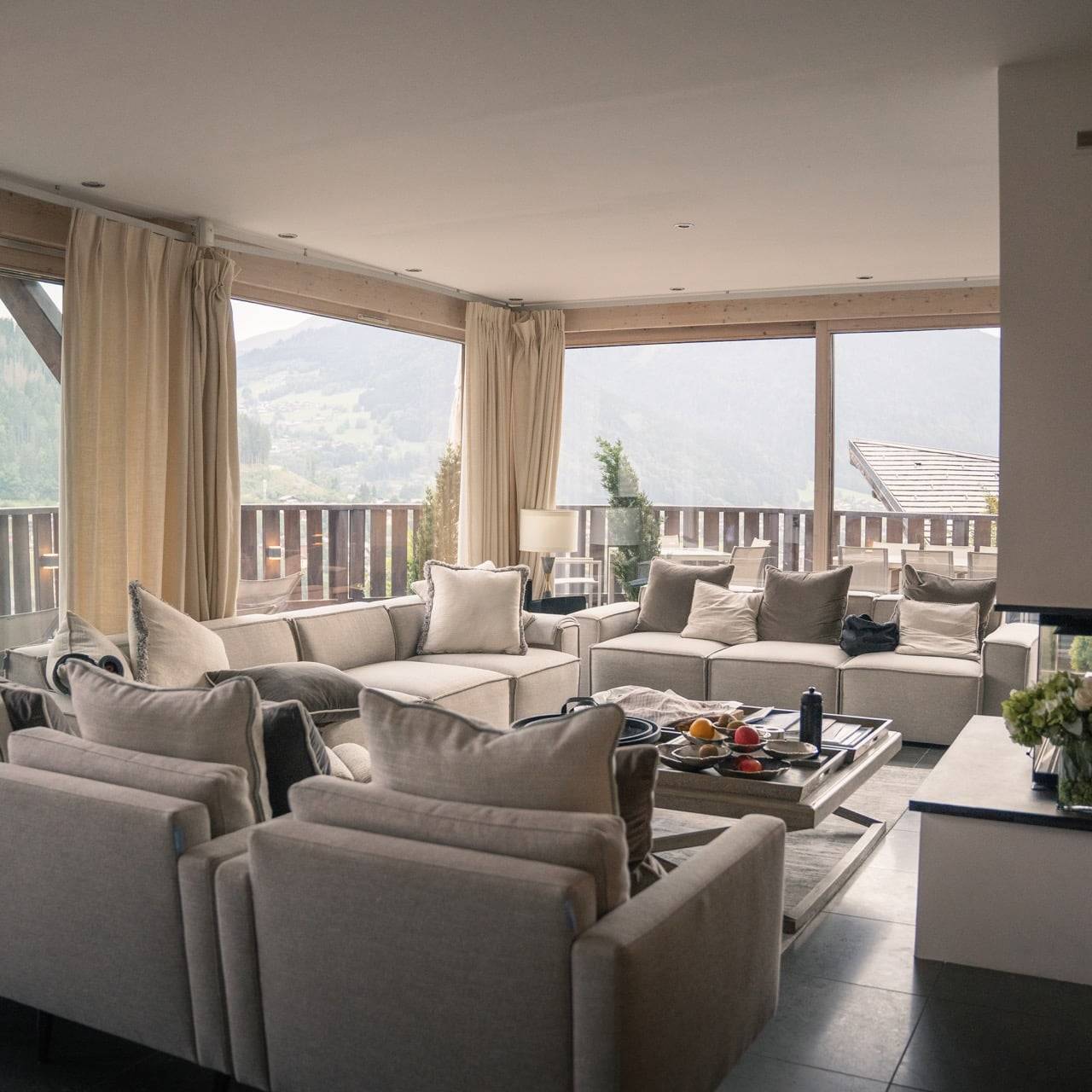 Spacious and cozy living room with beige sofas and armchairs arranged around a coffee table, set in a bright space with large windows showcasing scenic mountain views in the background.