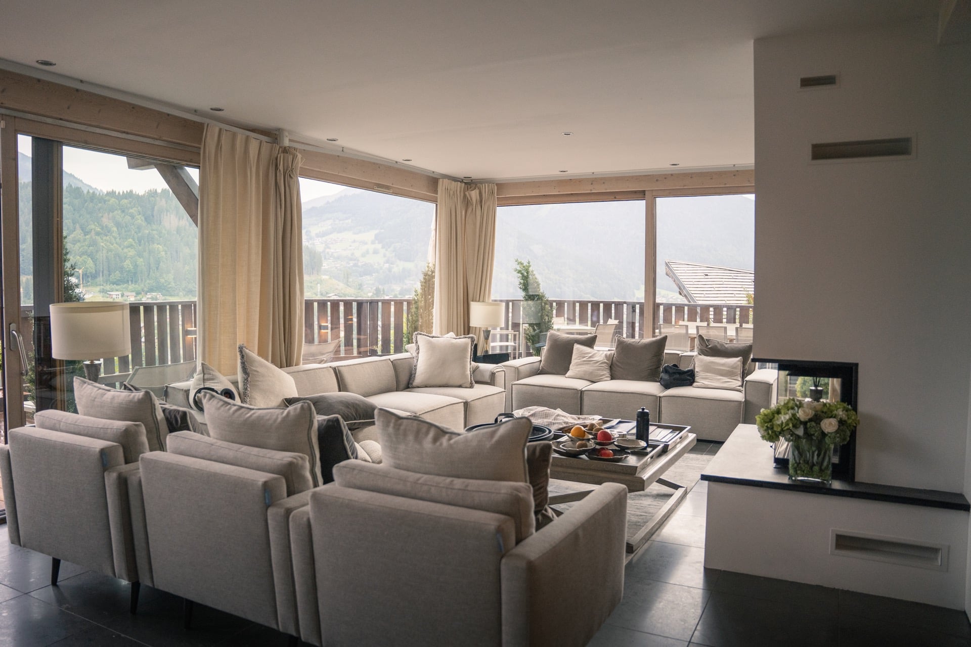 Spacious and cozy living room with beige sofas and armchairs arranged around a coffee table, set in a bright space with large windows showcasing scenic mountain views in the background.