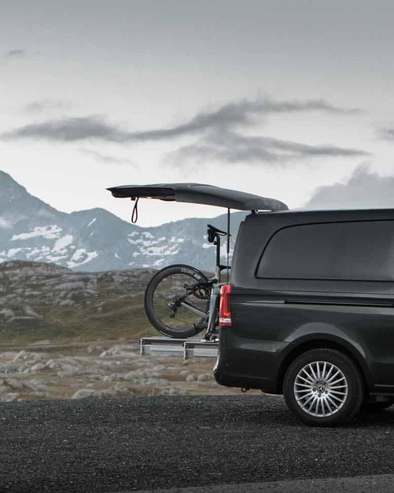 A sleek black van with its back door open, revealing a mounted bike against a backdrop of rugged mountains under a cloudy sky.