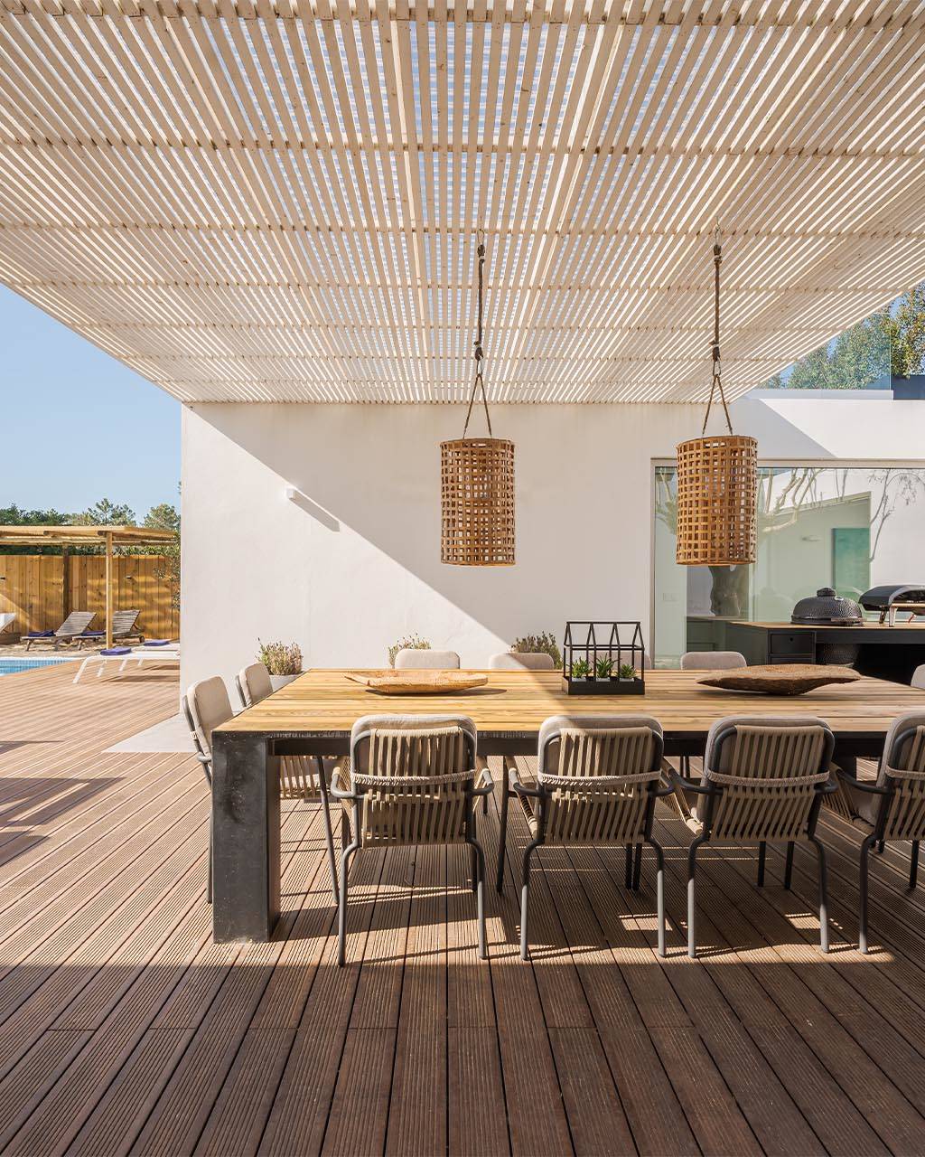 Modern outdoor patio with a wooden dining table, wicker pendant lights, and a view of a pool surrounded by lounge chairs and greenery.