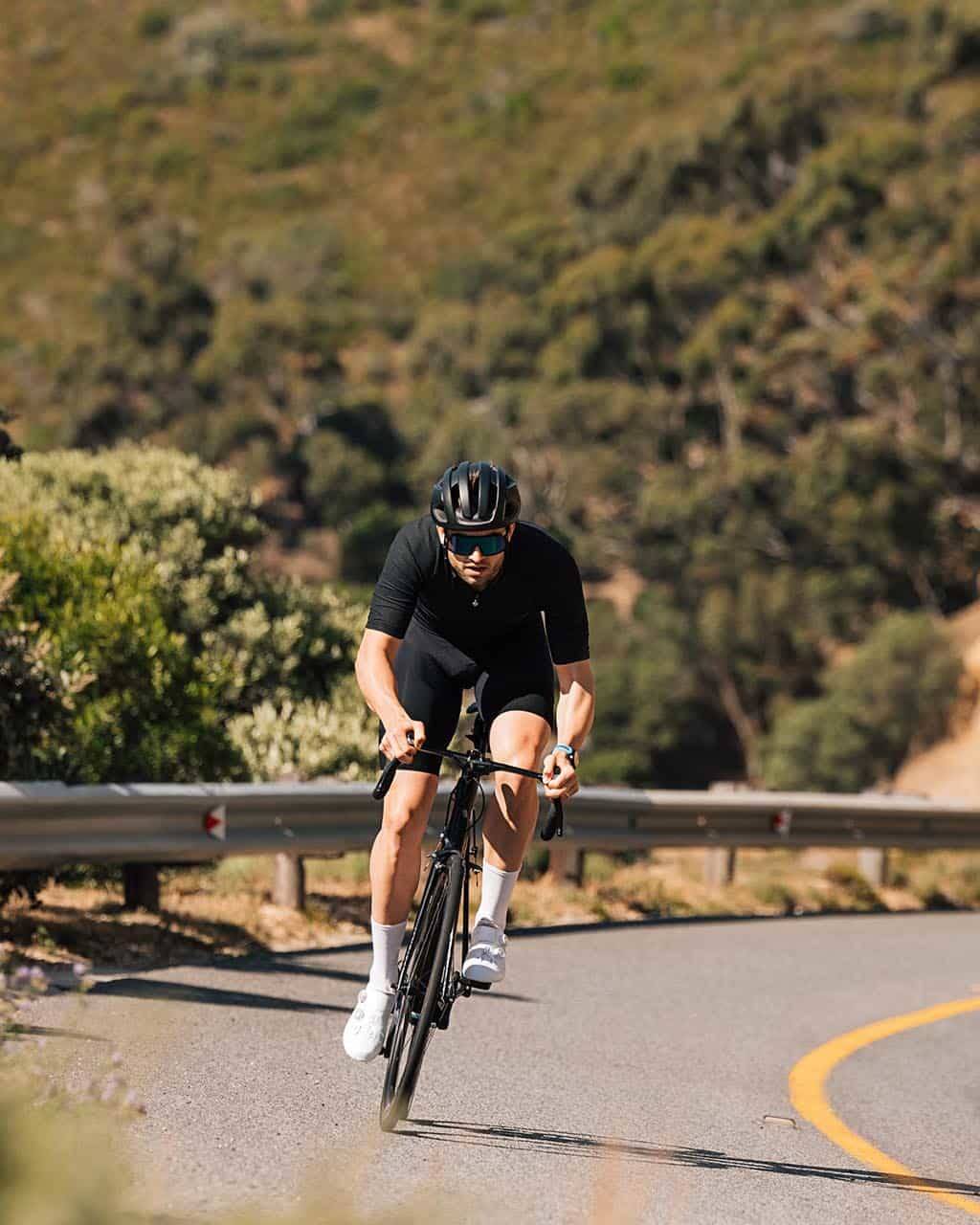 Focused cyclist dressed in black riding uphill on a curved mountain road, surrounded by greenery and rugged terrain.