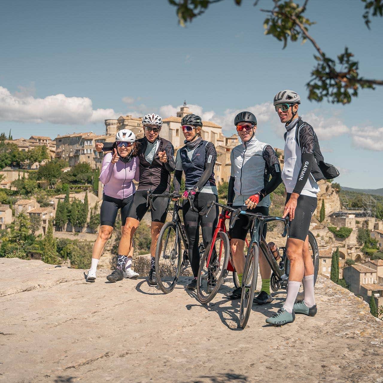 Group of five cyclists posing with their bikes on a sunny day, with a picturesque village and hills in the background.