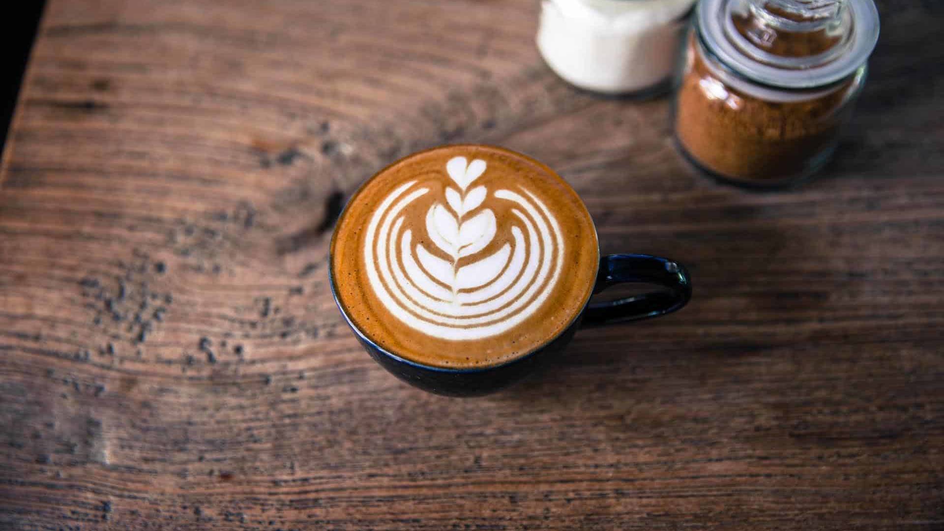 Close-up of a latte art design in a black cup placed on a rustic wooden table with jars of sugar and cinnamon in the background.