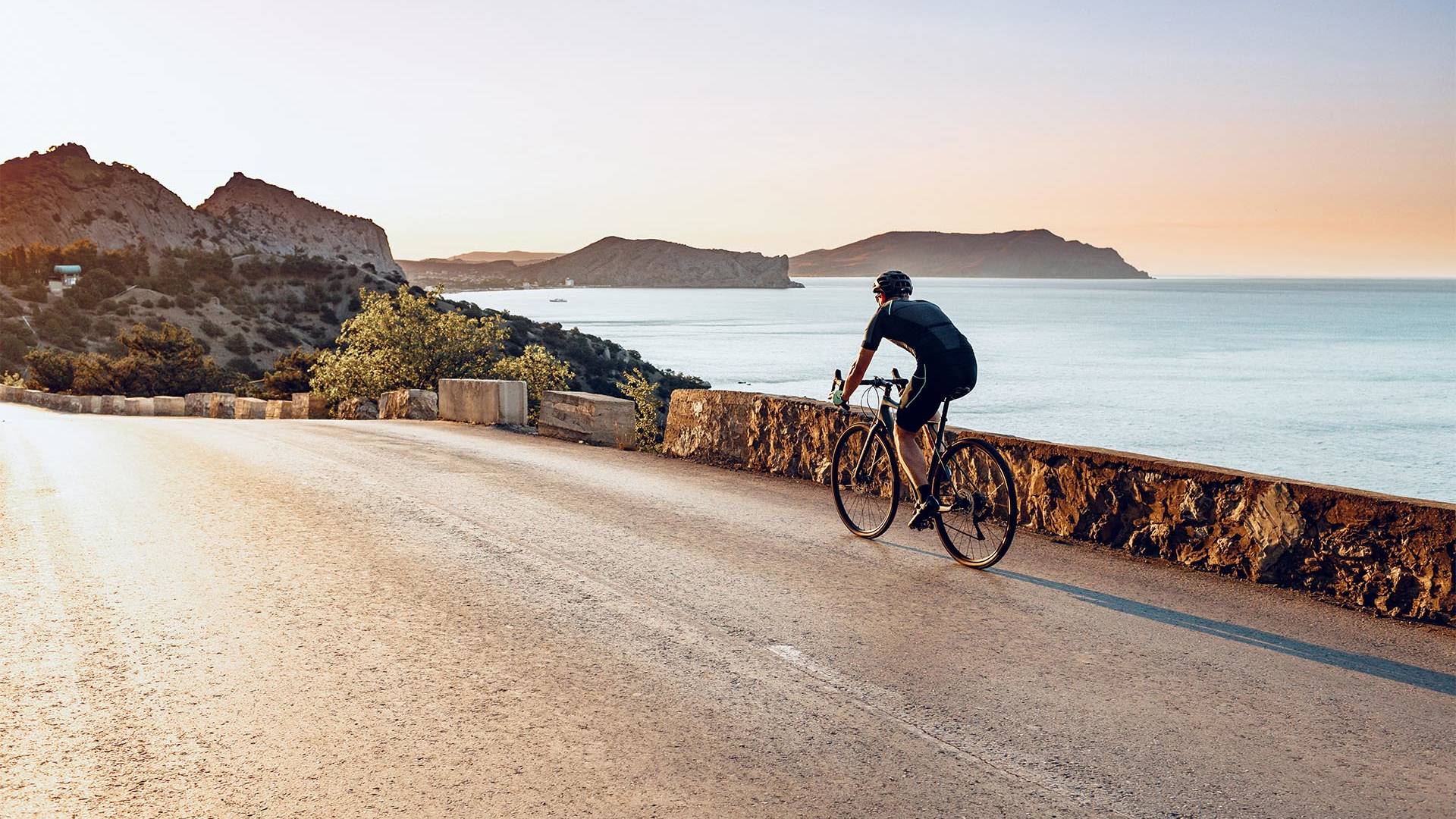 Cyclist riding along a coastal road during a serene sunset, with ocean views and rugged cliffs in the background.