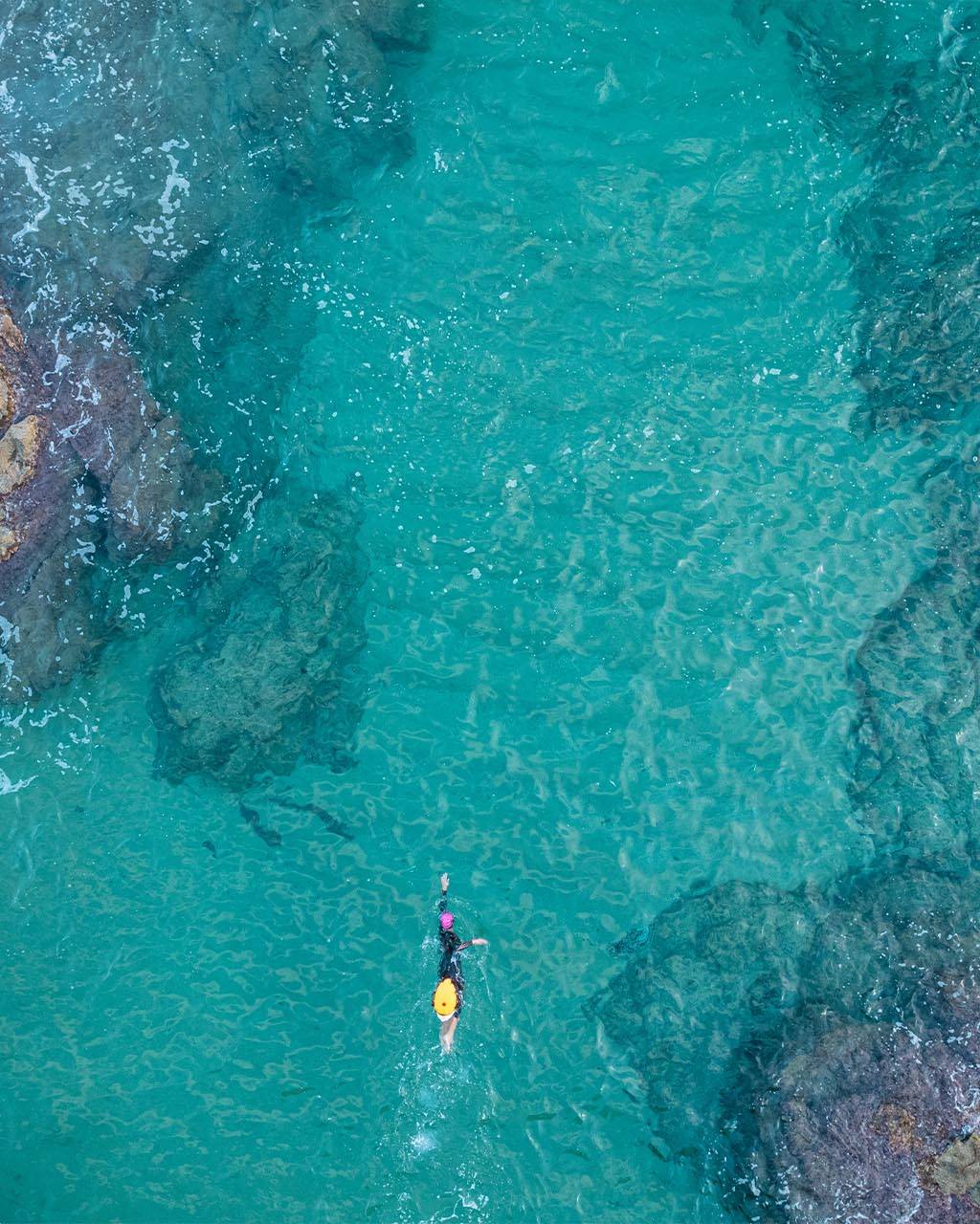 An aerial view of a swimmer in crystal-clear turquoise water near rocky shores, showcasing the ideal environment for learning how to choose the right triathlon camp for open-water training.