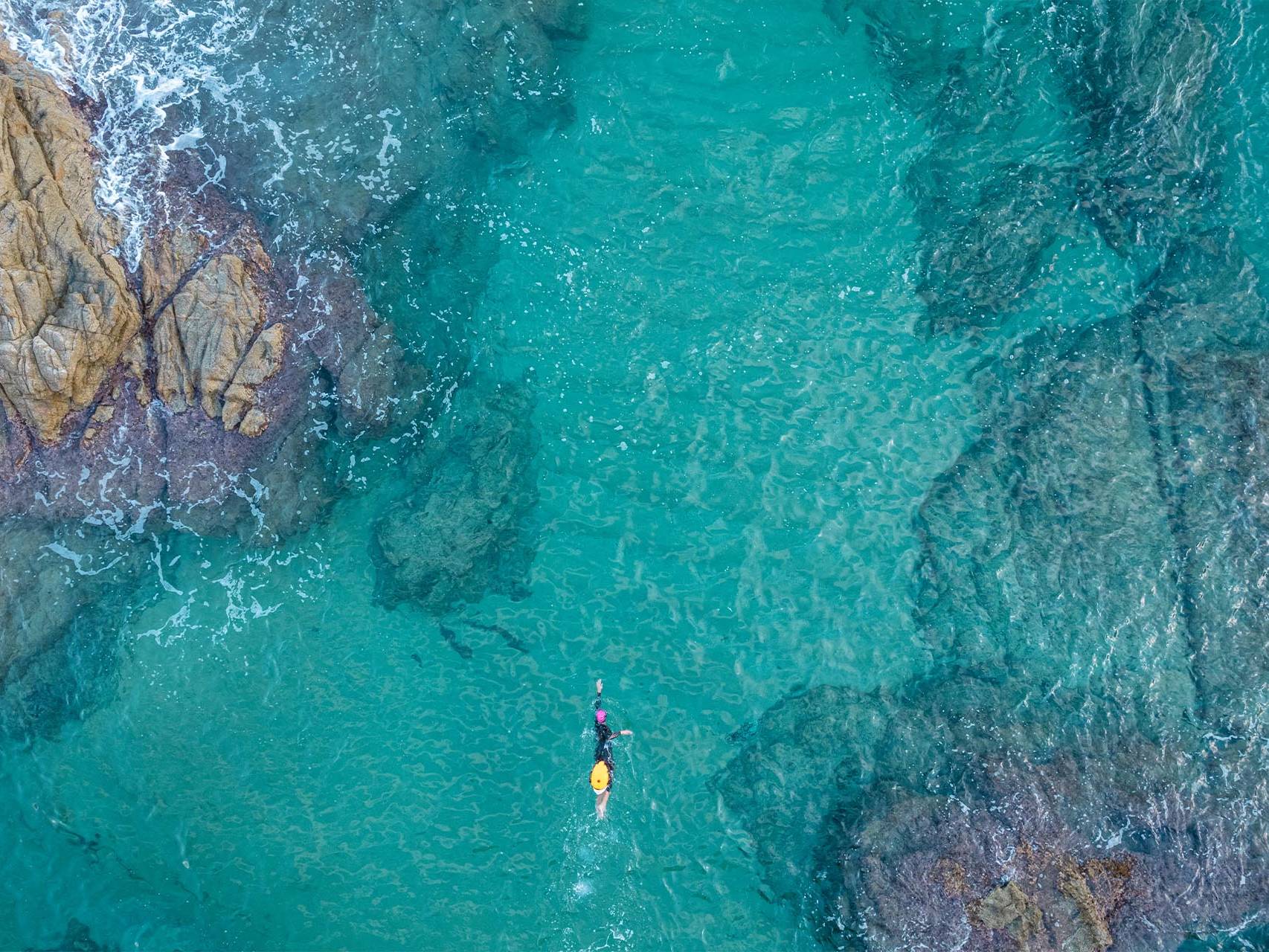 An aerial view of a swimmer in crystal-clear turquoise water near rocky shores, showcasing the ideal environment for learning how to choose the right triathlon camp for open-water training.